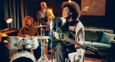 Mixed Race Woman Singing And Playing Guitar While Sitting On Chair With Legs Crossed. In Background Drummer, Saxophonist And Bass Guitarist.